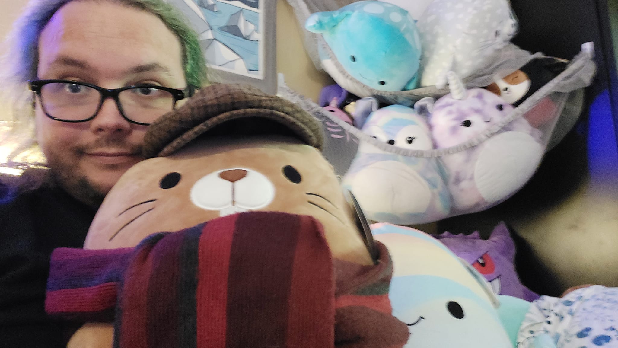 A man with long green hair wears pyjamas, holding a beaver Squishmallow dressed with his flat cap and red scarf, other Squishmallows are visible on the bed and in two tiers of hammocks